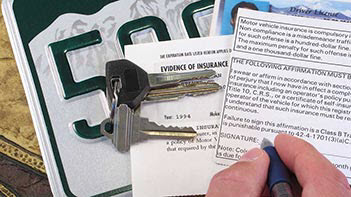 Photo of man signing auto registration form with a license plate, car title, evidence of insurance form and keys sitting by