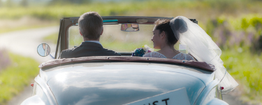 Couple in wedding attire driving into sunset in car with Just Married sign on it.