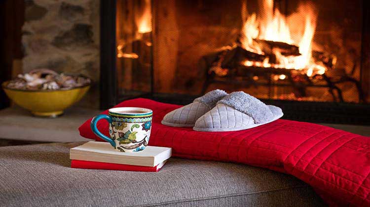 Libros, bebida caliente, cobija roja y pantuflas frente a la chimenea.