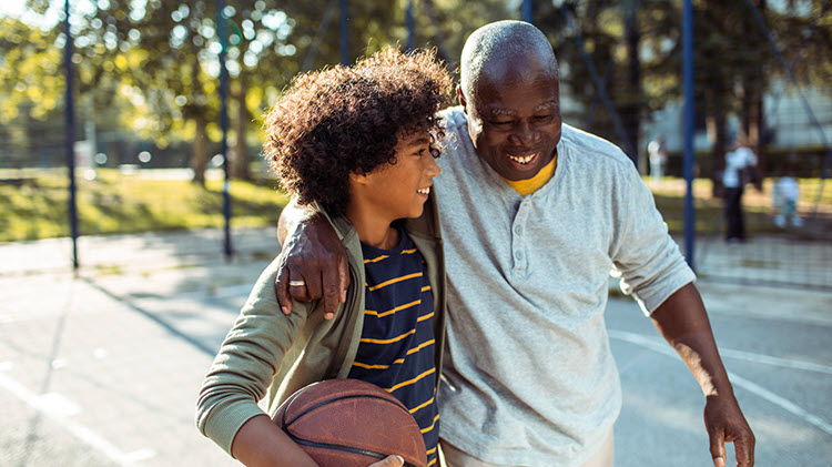 Boy carries a basketball and walks arm-in-arm with his grandfather across a basketball court.